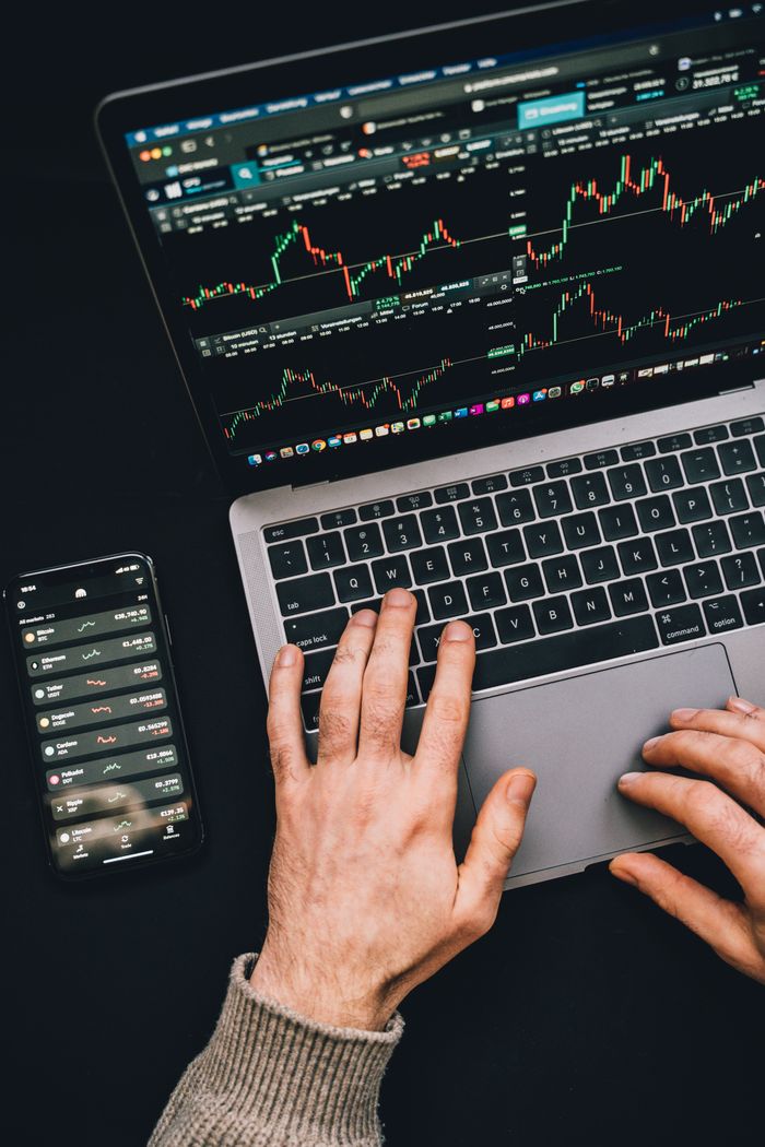 Mains en train de taper sur un ordinateur portable avec des graphiques de cryptomonnaie et l’écran d’un smartphone visibles, en mettant l’accent sur la finance numérique.